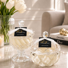 Two clear glass crystal look vessels with lids, filled with fragrant wax, displayed on a marble surface in a bright, softly lit interior. Each bottle has a black tag reading 'LV COLLECTION LUXURY FRAGRANCES' with a white ribbon.