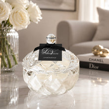 A clear faceted glass vessel with a black ribbon label reading LV COLLECTION LUXURY FRAGRANCES' sits on a marble table in a softly styled interior. White roses in a glass vase, a sofa, and a Chanel Dior book are visible in the background.