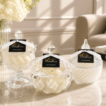 Three clear glass jars with lids and white ribbon bows are arranged on a marble surface in a bright interior setting. Each jar has a black tag reading 'LV Collection Luxury Fragrance.