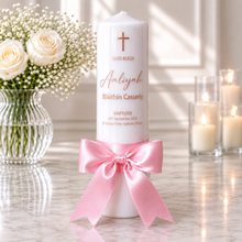 A personalized white pillar candle with a pink satin bow, featuring a printed brown cross and text for a baptism, styled on a marble surface with white roses and lit candles in the background.