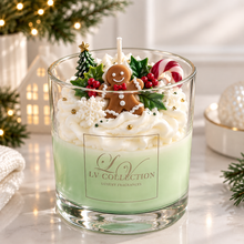 A festive holiday-themed scented candle in a clear glass jar from LV Collection, filled with pale green wax and topped with whipped cream, a gingerbread person decoration, a small Christmas tree, holly, red berries, and candy canes. The candle is set on a marble surface with warm holiday lights and greenery softly blurred in the background.