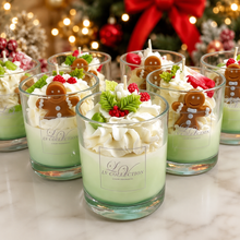 A set of holiday-themed scented candles in clear glass jars, decorated with whipped cream, green leaves, red berries, and gingerbread men, arranged on a marble surface with a festive Christmas tree in the background.