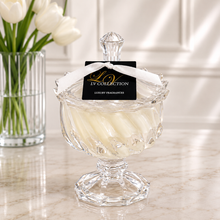 A clear glass pedestal jar with a faceted lid and a black hanging tag reading LV COLLECTION, filled with pale cream wax or candle-like strips, set on a marble surface with white tulips in a vase blurred in the background.