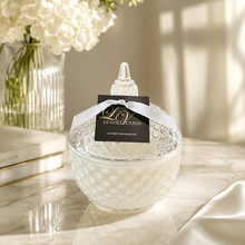 A clear textured glass jar with a matching lid, tied with a white ribbon and a black tag reading LV COLLECTION, set on a marble surface with soft sunlight and white flowers in the background.