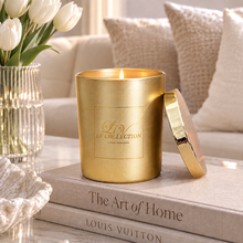 A gold metal scented candle jar with its lid leaning beside it, placed on stacked books in a softly styled interior setting. A vase of white tulips and neutral pillows are visible in the background.
